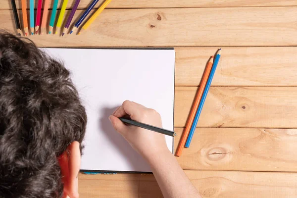 Child holding a pencil on top of a blank sheet surrounded by colored pencils