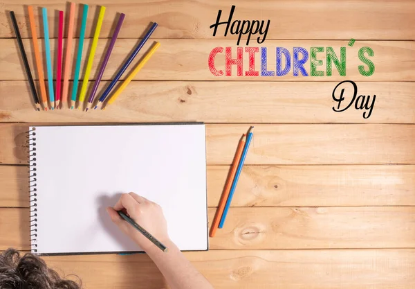 Child holding a pencil on top of a blank sheet surrounded by colored pencils. Text: Happy Childrens Day