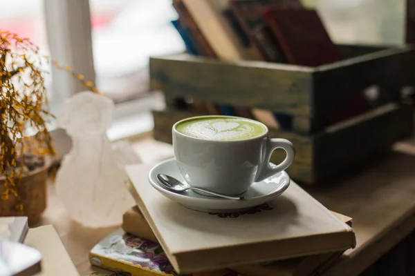Yeşil çay matcha latte sanat Kupası pencere yakınındaki kitaplarla ahşap masa üzerinde bir kahve dükkanı kafede bir açıyla kesilmiş. Arka plan.