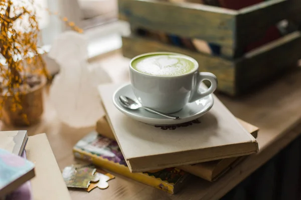 Yeşil çay matcha latte sanat Kupası pencere yakınındaki kitaplarla ahşap masa üzerinde bir kahve dükkanı kafede bir açıyla kesilmiş. Arka plan.