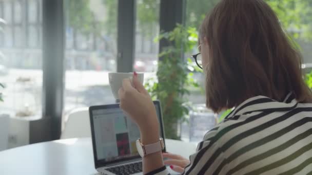 Za levým ramenem: dívka pracuje s notebookem, drží pohár — Stock video