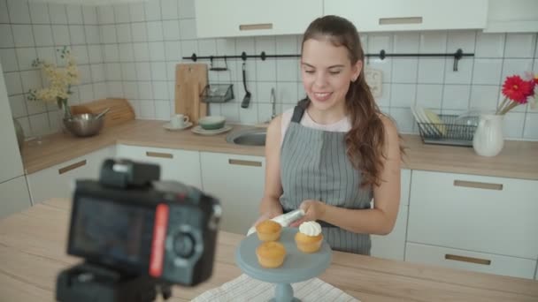 エプロンの美しい若いお菓子メーカーは ビデオ レッスン マスター クラス ビデオ Capcake の準備とクリーム カップ ケーキを飾るプロセスでを記録します — ストック動画