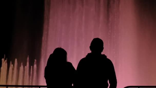 Silhouette of a pair of people standing against a colored fountain. — Stock Video