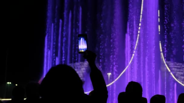 一个人的剪影在一个有色喷泉的背景下 举起他的手 拿着手机拍照或录像 彩色水显示喷泉 视频拍摄 游客拍摄视频 — 图库视频影像