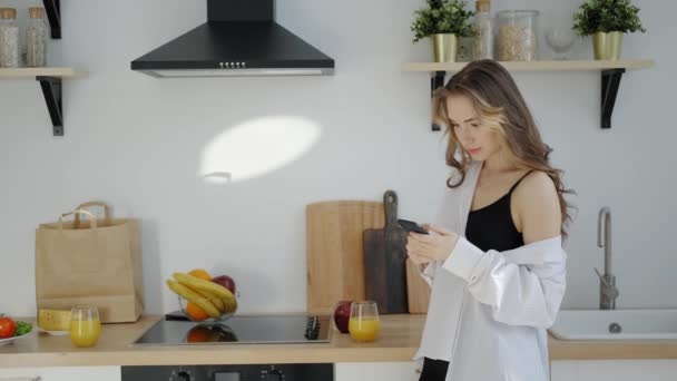 Jonge vrouw in wit herenshirt met smartphone in de ochtend keuken — Stockvideo