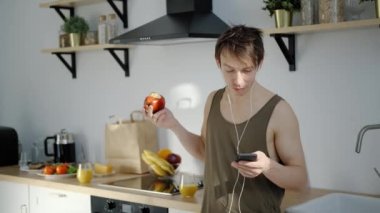 Neşeli genç adam elma yiyor, hafif mutfakta müzik dinliyor