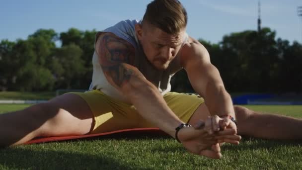 Tattooed muscular sportsman sitting on mat and bending forward to stretch leg muscles on green grass of athletic field — Stock Footage Tattooed Muscular Sportsman Sitting Mat Bending Forward Stretch Leg Muscles Royalty Free Stock Video