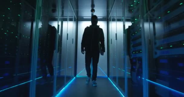 Хакер в маске, проходящий через ряды серверов — стоковое видео