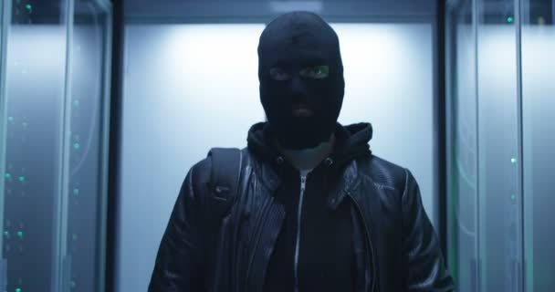 Хакер в маске, проходящий через ряды серверов — стоковое видео