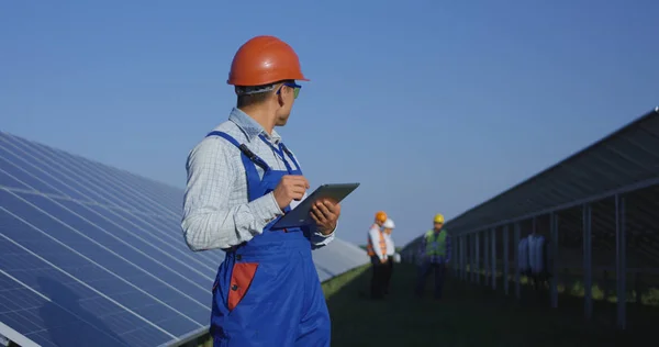 Trabalhador étnico em chapéu duro usando tablet entre painéis solares — Fotografia de Stock