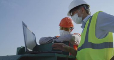 İnşaat alanında dizüstü bilgisayar kullanan maskeli mühendisler