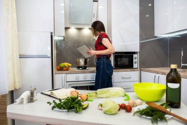 Sağlıklı beslenmeyle ilgili her şey için mutfağında farklı sebzeler pişirmek üzere tablet bilgisayar kullanan genç bir kadın.