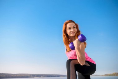 Formda kız, gölün kenarında, elinde dambıllarla egzersiz yapıyor.