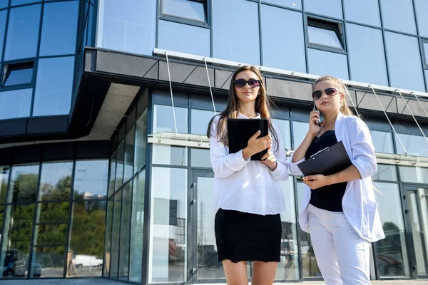 Two young business women with tablet and clipboard posing outside business center