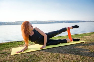  Genç bir kadın gündüz vakti göl kenarında direniş bandıyla egzersiz yapıyor. Sağlıklı yaşam tarzı