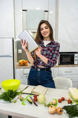 Sağlıklı beslenmeyle ilgili her şey için mutfağında farklı sebzeler pişirmek üzere tablet bilgisayar kullanan genç bir kadın.