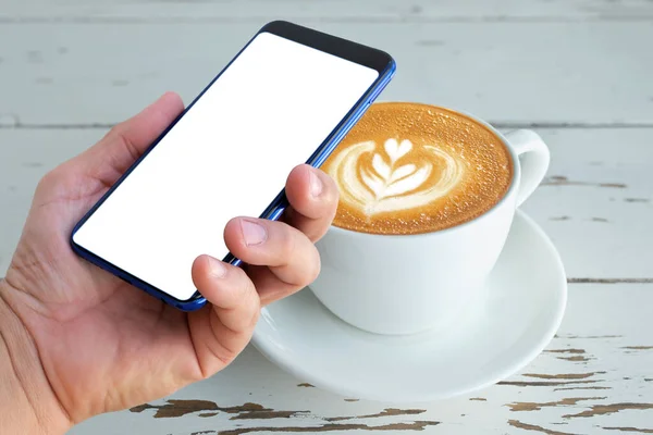 Boş ekranlı akıllı telefonu latte kahve üzerine tut. Kalbini beyaz bardakla süsle, ahşap masada.