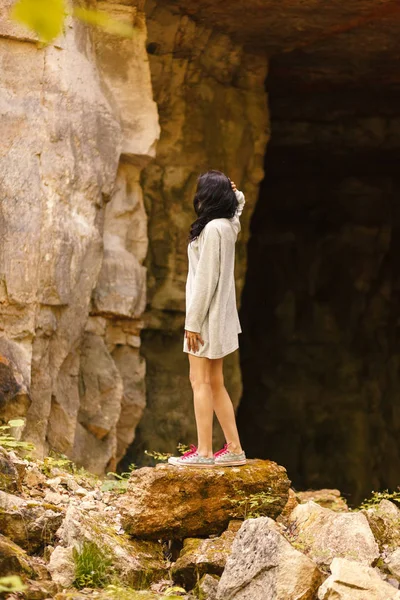 A woman in a long t-shirt and sneakers with pink laces stands on a large stone on the background of high rocks. Woman standing in front of cave entrance