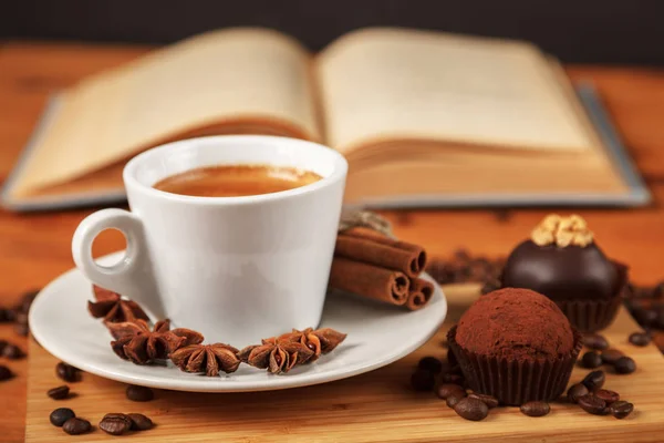 Coffee break a Cup of hot coffee and cakes on a wooden table with an open book. A white Cup of black coffee surrounded by a small amount of roasted coffee beans, cinnamon and anise