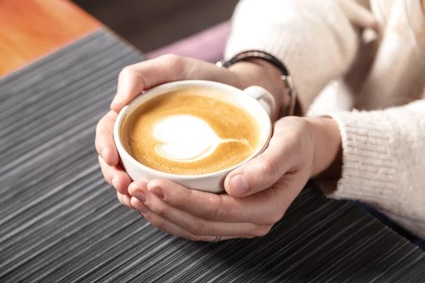 Yakın çekim sıcak latte sanat kalp şeklinde bir genç kızın elinde kahve beyaz bir fincan. Sıcak kış örme beyaz kazak giyen kadın. Tonda. Kahve kız kavramı gevşeme kahve kupa.