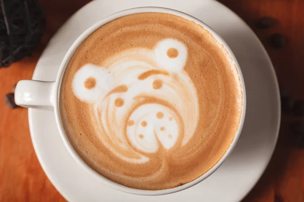 White Cup cappuccino coffee with a latte-art pattern in the form of a bear close-up on a wooden background. The concept of making coffee in a coffee shop