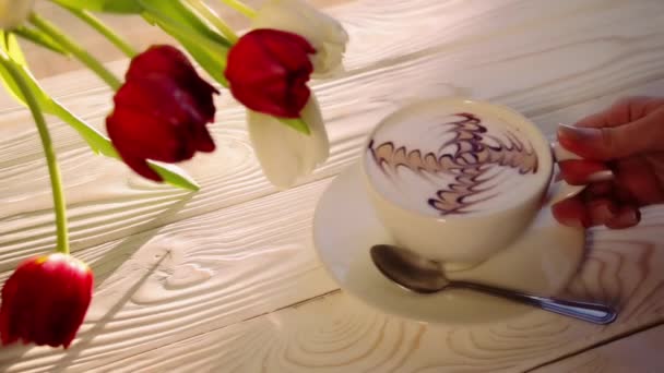 拿着一杯咖啡 上面有拿铁艺术图案 放在一张白色的木桌上 上面有一束郁金香 — 图库视频影像