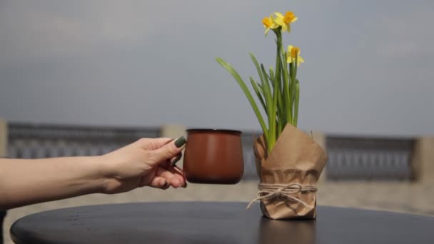 ウォーターフロントのオープンカフェで 水仙の花束を持つテーブルの上の朝のコーヒー 人々はぼやけた背景を通り過ぎる — ストック動画