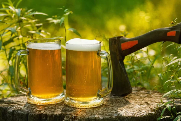 Beer mugs cheers, picnic or party on natural background with axe.