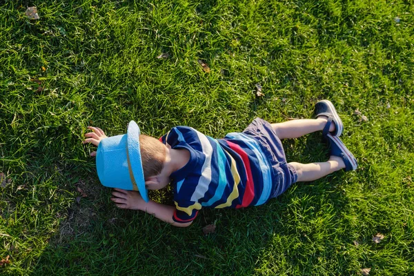 Un niño guapo tirado en la hierba. El pequeño soñador yace en el prado con sombrero. Niño en la naturaleza fuera de la ciudad. Vacaciones sin preocupaciones —  Fotos de Stock