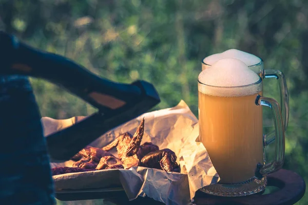 Glass of light beer against background of green trees outdoor. Snack sausage, smoked chicken wings, vintage toning picture — Stock Photo, Image