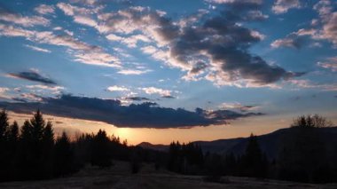 Gün batımının alacakaranlığa dönüştüğünü gösteren bir zaman çizelgesi. Mavi gökyüzünde renkli bulutlar, ufukta kaybolan güneş ışınları ve ormandaki ağaçların silueti. Ön plandaki çayır.