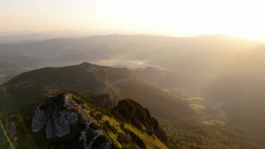 Bir hava videosu, heybetli bir dağ zirvesi ve çevresini gösteren yavaş bir doli hareketini gösteriyor. Sahne, batan güneşin yumuşak ışığıyla aydınlanır.. 