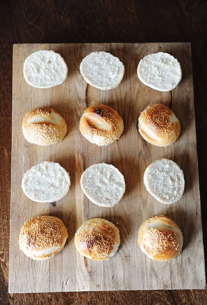 Bollos de hamburguesas de sésamo divididos por la mitad en la vista superior de tabla de cortar de madera. Proceso de cocción —  Fotos de Stock