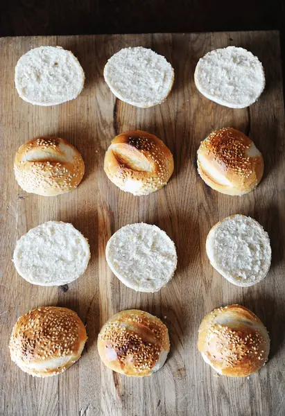 Bollos de hamburguesas de sésamo divididos por la mitad en la vista superior de tabla de cortar de madera. Proceso de cocción —  Fotos de Stock