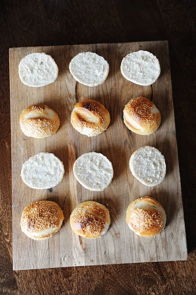 Bollos de hamburguesas de sésamo divididos por la mitad en la vista superior de tabla de cortar de madera. Proceso de cocción —  Fotos de Stock