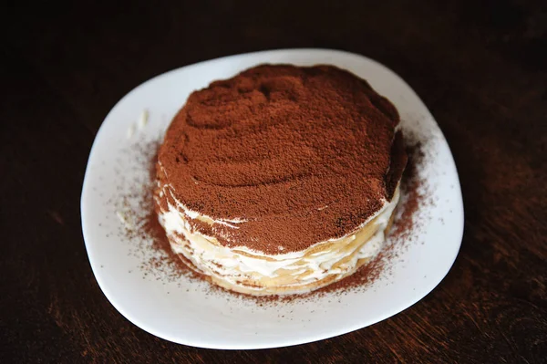 Свежеприготовленный торт из тирамису, посыпанный порошкообразным какао. Белая тарелка, деревянный стол — стоковое фото