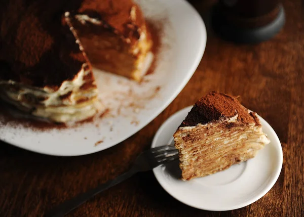 Piece of crepe cake with tiramisu filling and powdery cocoa is served on plate. The interior of coffee shop