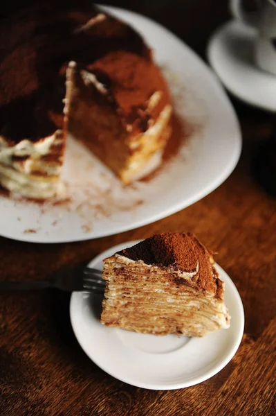 Un pedazo de pastel de crepe con relleno de tiramisú y cacao en polvo se sirve en el plato. El interior de la cafetería —  Fotos de Stock