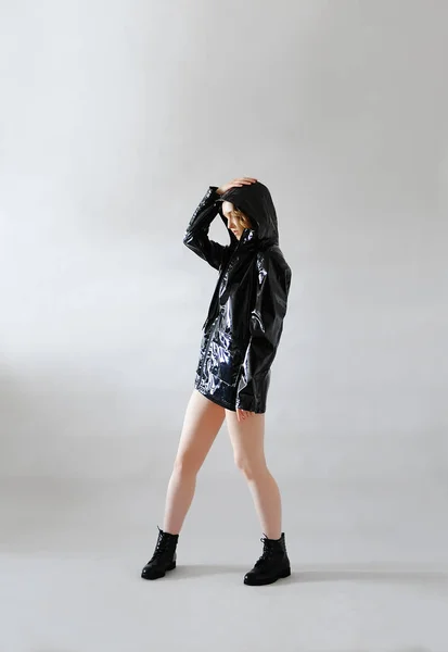Stylish young girl in black lacquered rain jacket raincoat with hood and low shoes. Light background. Trendy outfit