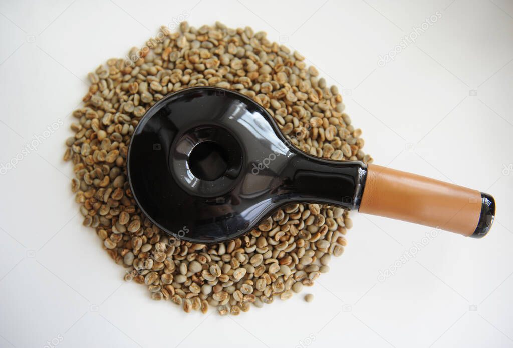 Home coffee roasting. Manual black ceramic sample roaster tiegel with leather handle on scattering of green coffee beans in the shape of a circle. White background