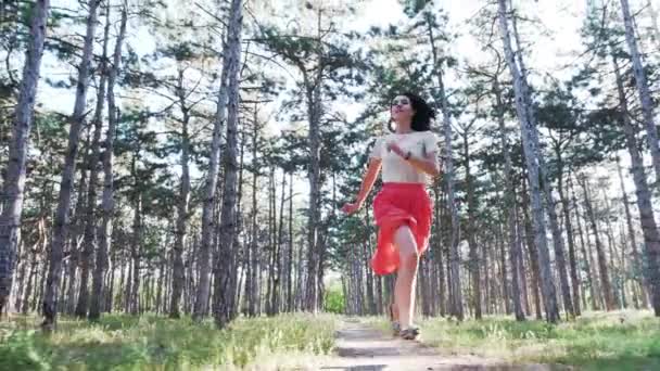 Uma jovem mulher bonita está feliz correndo ao longo da estrada entre numerosos pinheiros. Movimento lento — Vídeo de Stock