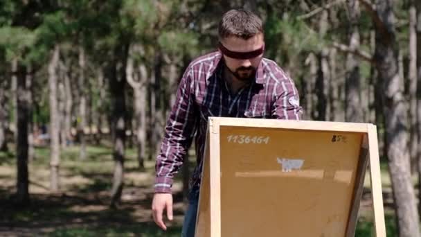 A blondfolded man enthusiastically makes sketches on a canvas while being in the woods — Stock Video