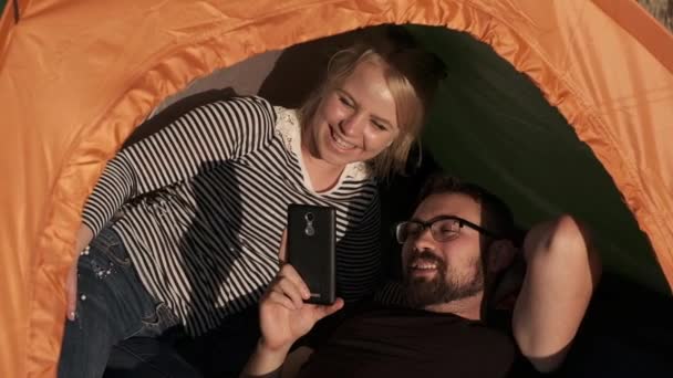 Una pareja feliz en una tienda de campaña está viendo algo por teléfono y riéndose — Vídeos de Stock