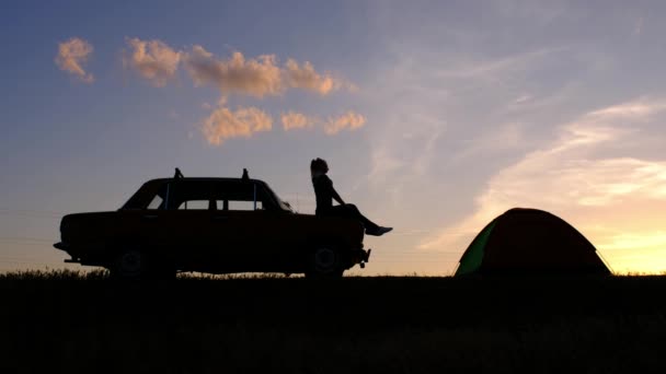 Uma silhueta de uma jovem mulher, sentada em um carro e fixando seu cabelo contra o pôr do sol — Vídeo de Stock