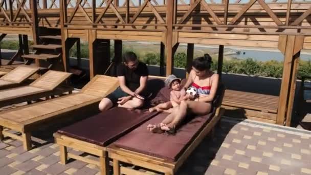 Blick auf eine Familie, die auf Chaiselongue vor dem Schwimmbad liegt und mit Fußball spielt — Stockvideo