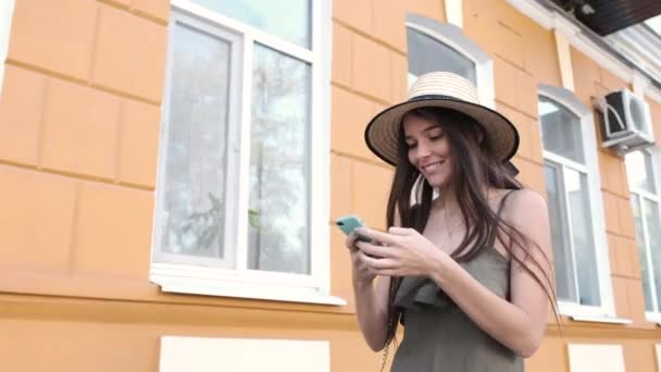 Una joven con un sombrero y un vestido largo está enviando mensajes de texto felices en su teléfono — Vídeos de Stock