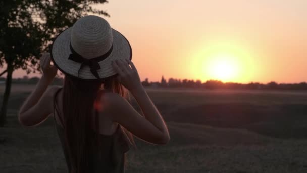 Uma jovem mulher está olhando para o pôr do sol enquanto segura seu chapéu — Vídeo de Stock