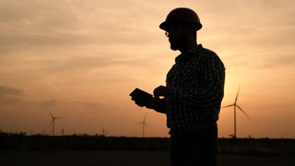 Silhueta de um engenheiro de usinas eólicas com um tablet nas mãos sobre um fundo de pôr do sol . — Vídeo de Stock