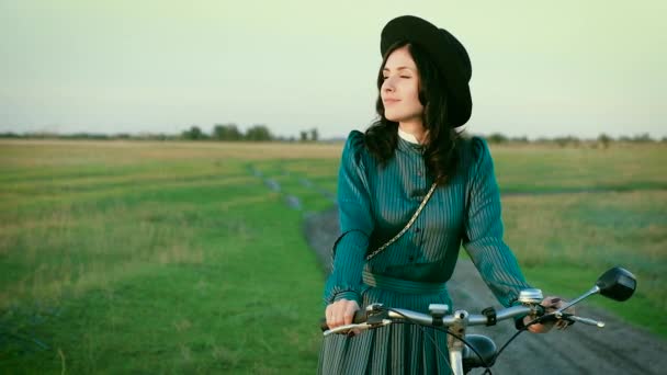 Retrato de una morena con sombrero en un vestido vintage y con una bicicleta en la estepa . — Vídeo de stock