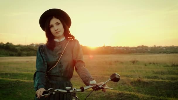Porträt eines Mädchens mit Hut in einem Vintage-Kleid und mit einem Fahrrad vor dem Hintergrund der Sonnenuntergangssonne. — Stockvideo